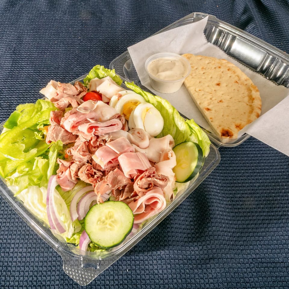 A chef salad with ham, turkey, hard-boiled eggs, and vegetables, with pita bread and dressing.