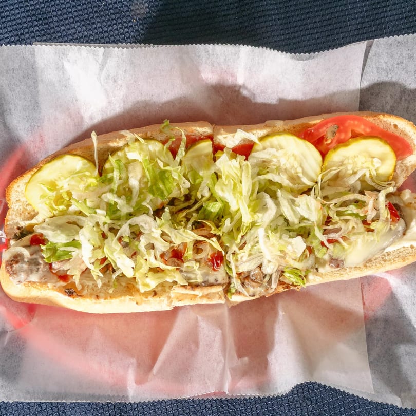 A Zeda's Pizza sub sandwich with lettuce, pickles, tomatoes, and melted cheese on a red tray.