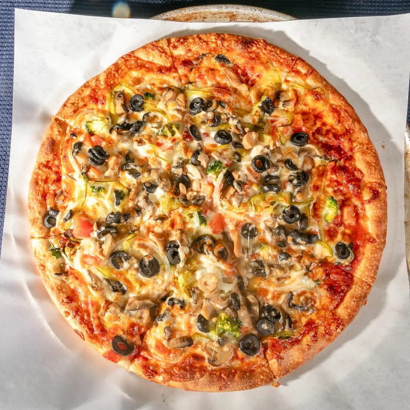 A whole Zeda's Pizza with black olives, mushrooms, onions, and peppers on parchment paper.